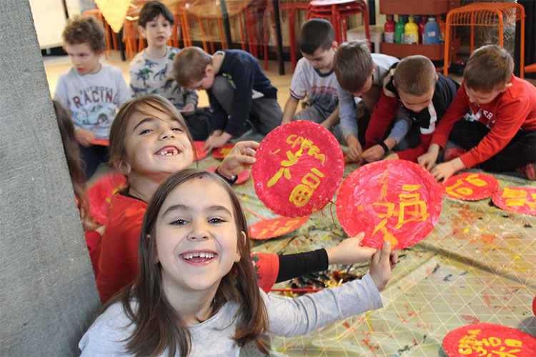 Deca sede na podu i pise kineske znakove zutom bojom na crvenoj podlozi u vrticu Povratak prirodi
