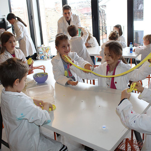 Deca i nastavnik u laboratorijskim mantilima rukama razvlace zutu elasticnu masu