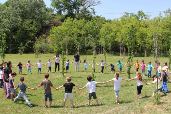 Vise desetina dece i nekoliko nastavnika drzeci se rukama formiraju veliki krug na livadi sa drvecem u pozadini